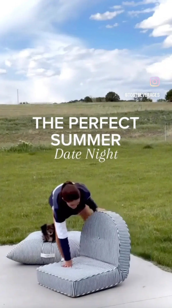 Person setting up striped outdoor lounge furniture for a perfect summer date night, with a small dog on a cushion in the background.