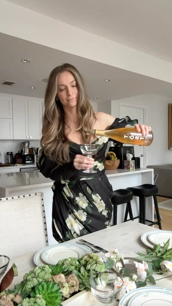 Person in a floral dress pouring TOST sparkling drink from a bottle into a glass on a beautifully set dining table, with elegant floral decorations and a modern kitchen in the background.