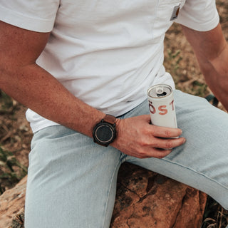 Person sitting outdoors holding a TÖST can, wearing a white shirt and light blue pants.

