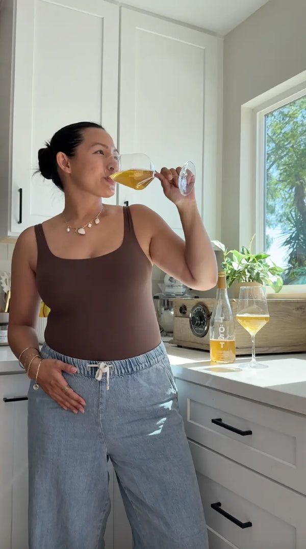 Person enjoying a glass of TOST sparkling beverage in a bright kitchen, wearing a brown tank top and casual jeans, with a bottle of TOST beverage and a second glass of drink on the counter.