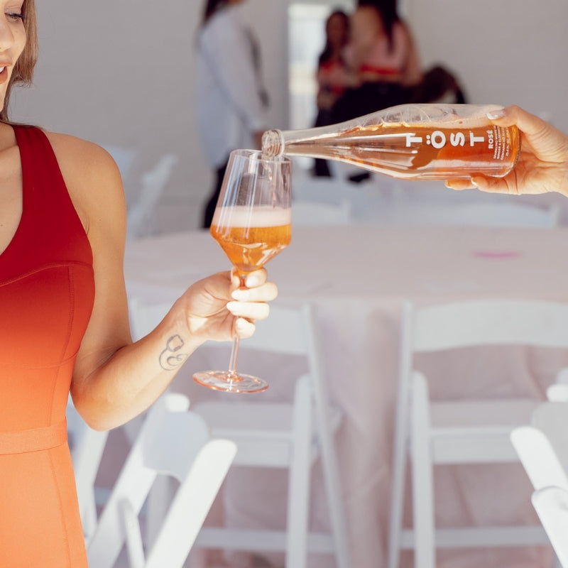 Person pouring a drink from a bottle labeled TOST into a glass, with a blurred indoor setting.
