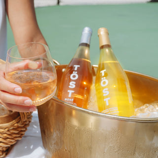 Two bottles of Töst wine alternative in an ice bucket with a glass of TOST held by a person.
