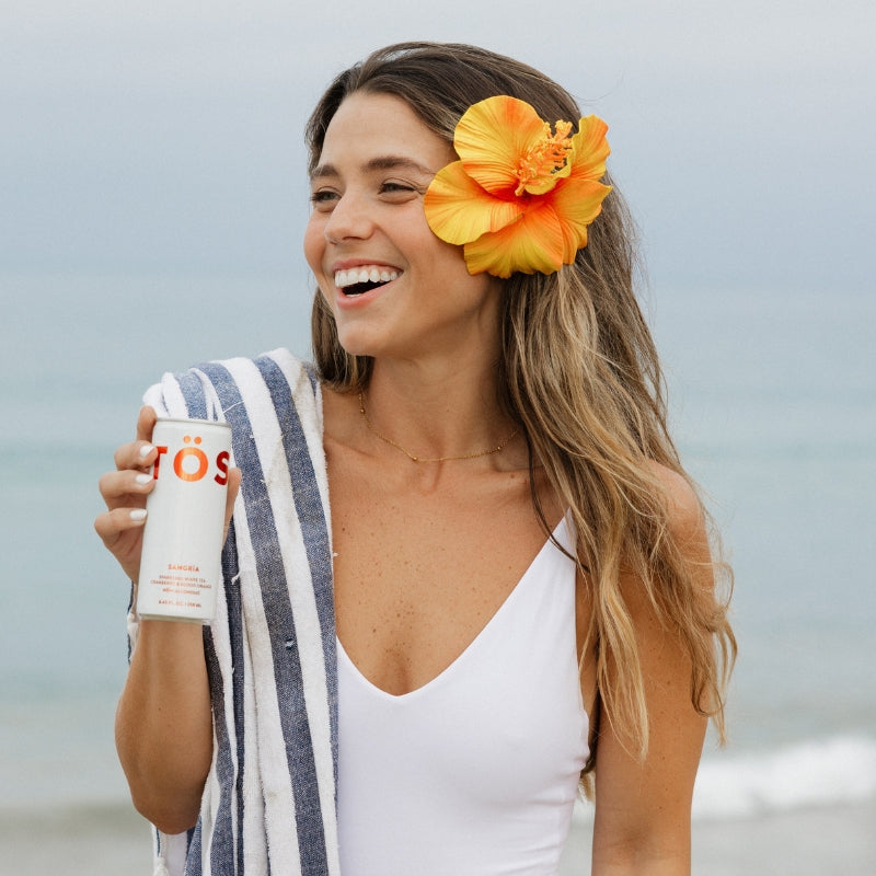 Woman on a beach holding a bottle of TOS with an orange flower in her hair.
