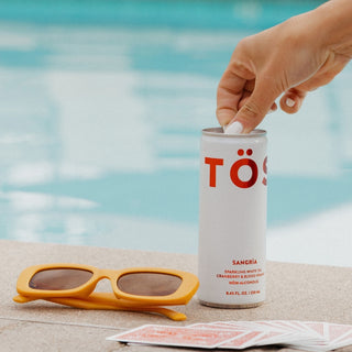 Hand opening a can of TÖST Sangria by a poolside with sunglasses and a deck of cards.
