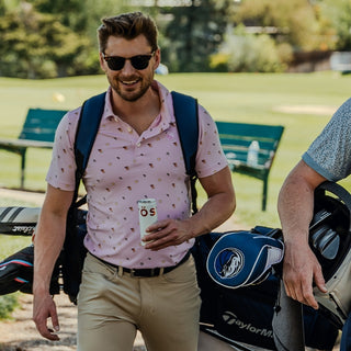 Man walking on a golf course holding a drink, wearing sunglasses and a patterned shirt.
