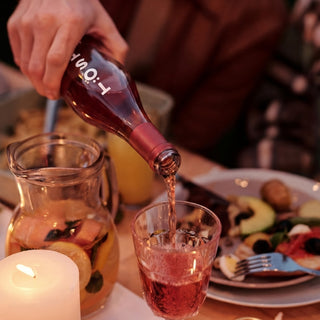 Person pouring TOST Sangria from a bottle into a glass on a dining table with food and a candle.
