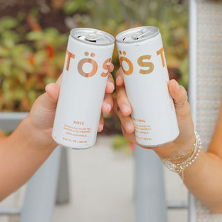 Two hands holding TOST cans in a celebratory manner with a blurred outdoor background.
