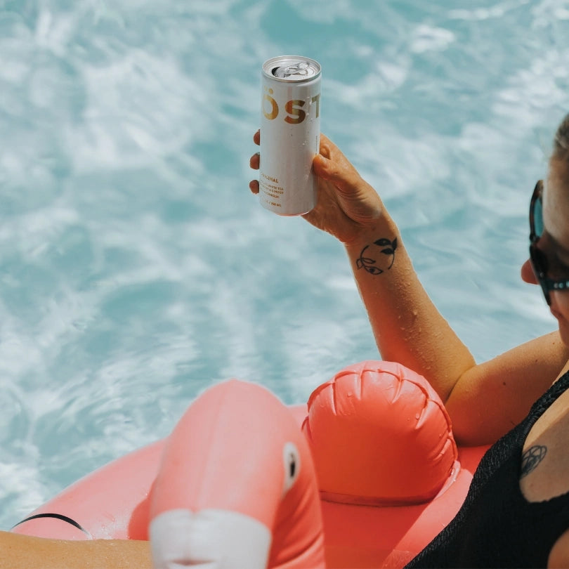 Person relaxing in a pool holding a can of TÖST non alcoholic sparkling beverage while floating on a pink inflatable, with the brand name “TÖST” visible against blue water.
